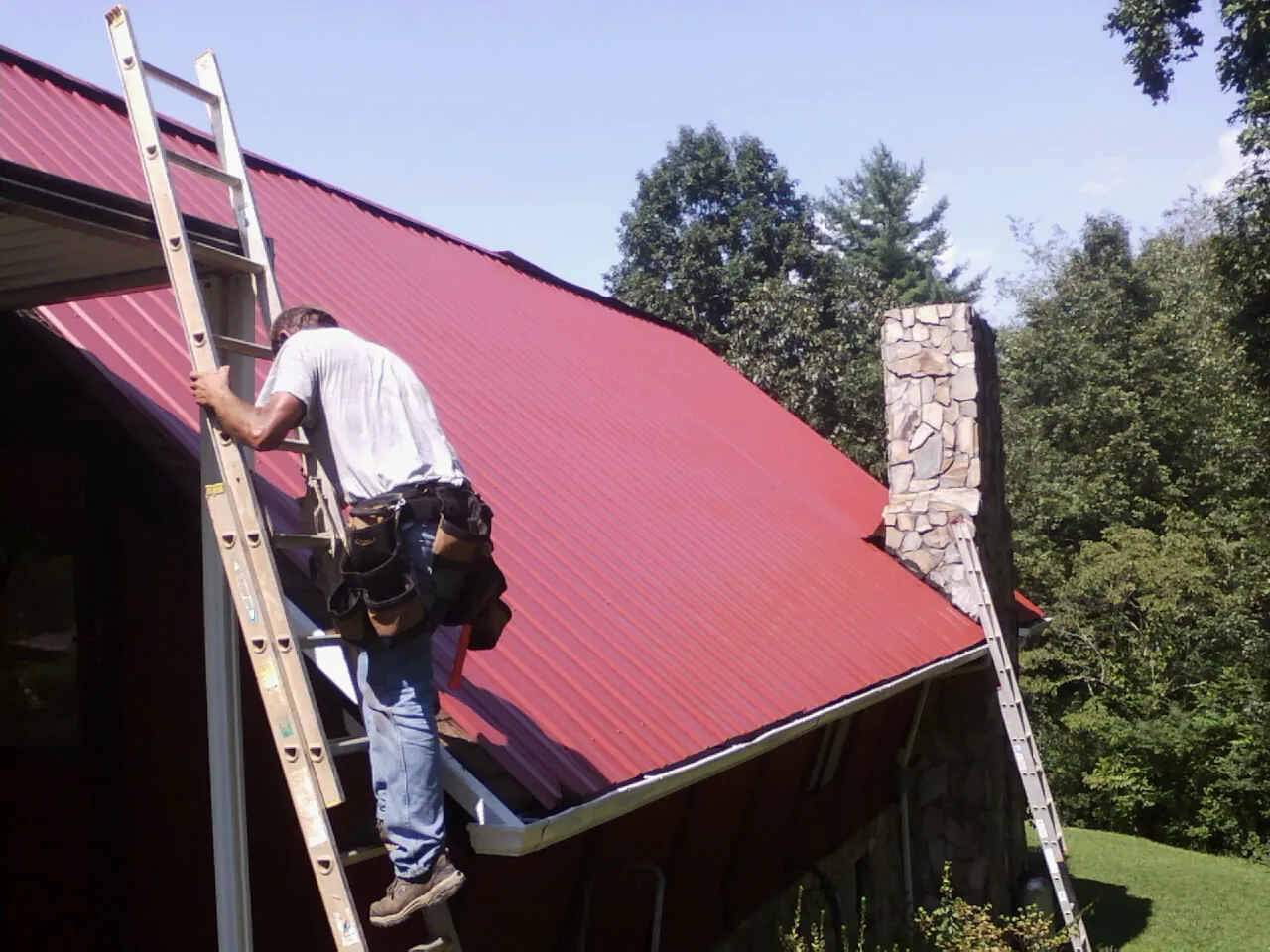 Roofer on a ladder beside a red metal roof and stone chimney — Soffit & Fascia Repair by Roofing Brothers in Sherborn